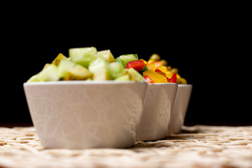 Vegetables in white bowls, diced on a brown kitchen table. Peppers, cucumbers, sweet corn and peas in a bowl on the table.