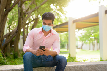 Obraz premium Young Indian businessman with mask using phone and sitting at the park