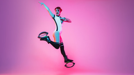 Fototapeta na wymiar Beautiful redhead woman in sportswear jumping in a kangoo jumps shoes isolated on purple-pink gradient studio background in neon light. Active movement, action, fitness and wellness. Fit female model.