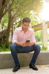 Fototapeta premium Full body shot of young handsome Indian businessman using phone and sitting at the park