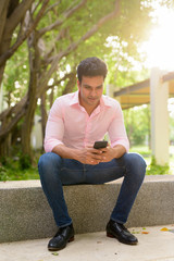 Obraz premium Full body shot of happy young handsome Indian businessman using phone and sitting at the park