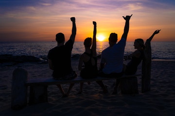 Group of four friends sitting on a wooden bench on the beach at sunset. They are with their hands up. The photo is taken from the back.