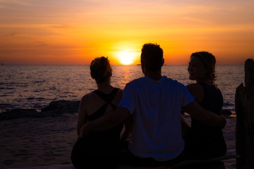 A boy and two girls sitting on a wooden bench on the beach at sunset. He's holding them by the waist.