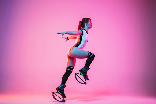 Beautiful redhead woman in sportswear jumping in a kangoo jumps shoes isolated on purple-pink gradient studio background in neon light. Active movement, action, fitness and wellness. Fit female model.