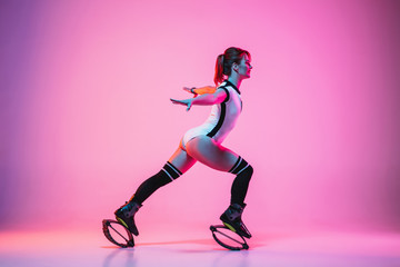 Beautiful redhead woman in sportswear jumping in a kangoo jumps shoes isolated on purple-pink gradient studio background in neon light. Active movement, action, fitness and wellness. Fit female model.