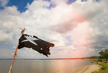 Black torn pirate flag Jolly Roger with crossed sabers waving on a stick against the background of the river and cloudy sky