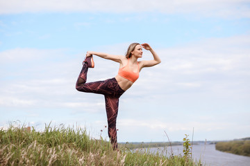 Attractive sportswoman is practicing yoga on the river bank. Dancer pose. Woman is stretching in Natarajasana exercise. Caucasian woman is wearing sportswear and sneakers. 
