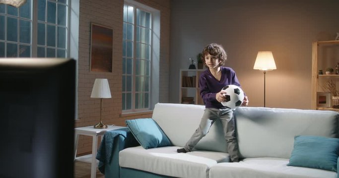 Funny expressive little asian boy watching a soccer game on tv and jumping on couch in front of it, reacting to his favourite team playing - football fans concept 4k footage