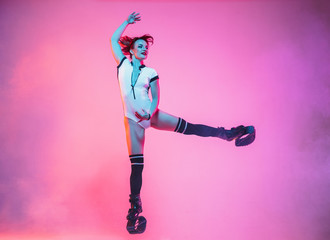 Beautiful redhead woman in sportswear jumping in a kangoo jumps shoes isolated on purple-pink gradient studio background in neon light. Active movement, action, fitness and wellness. Fit female model.