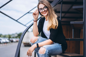 Obraz na płótnie Canvas Beautiful girl with long hair and glasses sitting on metal stairs on the wooden background of house with vertical boards. Woman smiling and looking at camera