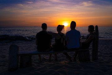 Group of four friends sitting on a wooden bench on the beach at sunset. In the background you can see the sun. From the back.