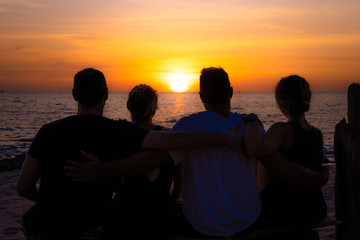 Four friends hugging watching the sunset. They are on the beach sitting on a wooden bench from the back. In the background you can see the sun and the sea.