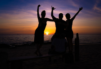 Group of four friends at sunset on the beach. One boy is sitting on the shoulders of another and the girls are standing on a wooden bench. The sun looks between them.