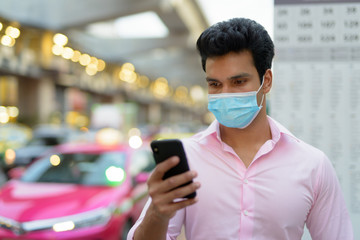 Face of young Indian businessman with mask using phone at the bus stop