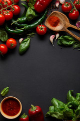 top view of red cherry tomatoes, tomato sauce in bowl, peppercorns, herbs and green chili peppers on black