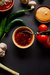 top view of tasty sauces in bowls near ripe cherry tomatoes, garlic, green chili peppers and mushrooms on black