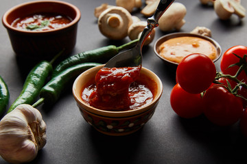 selective focus of spoon in tasty tomato sauce near ripe vegetables on black