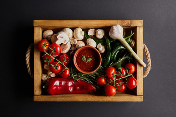 top view of tomato sauce with basil leaves near cherry tomatoes, green chili peppers, mushrooms and rosemary in wooden box on black