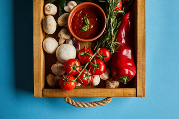 top view of tomato sauce in bowl on mushrooms near red cherry tomatoes, rosemary and chili pepper in wooden box on blue