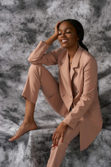 Elegant african woman in pink costume. Fashion studio portrait. Female model in trousers and blazer. Lady sitting and posing at gray background.