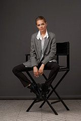 Male style dressed fashion female model. Fashionable woman in blouse, blazer, trousers and boots sitting at high cjair and posing against gray background. Stylish fashionable blonde girl