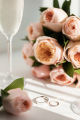 Wedding rings lie next to two glasses of champagne and a bouquet of roses in the sun