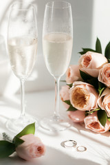 Wedding rings lie next to two glasses of champagne and a bouquet of roses in the sun