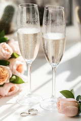 Wedding rings lie next to two glasses of champagne and a bouquet of roses in the sun