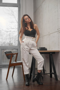 Portrait with soft light of elegant beautiful tanned caucasian woman with long straight brunette woman in black lace top and white trousers posing in loft interior wth concrete walls and big window