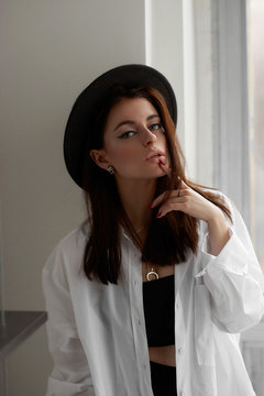 Elegant beautiful slim tanned caucasian woman in black top, leggins, white oversize shirt and hat standing near wall in bright loft interior and posing. Fashionable trenndy stylish female