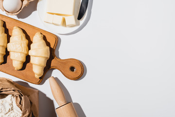 top view of fresh croissants on wooden cutting board near flour, rolling pin, butter and egg on white background