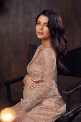 Young beautiful elegant pregnant woman wearing luxury expensive evening dress, sitting at high chair and posing alone in dark stylish interior