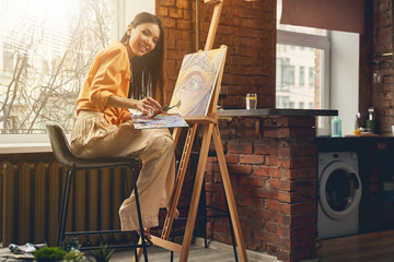 Joyful young woman painting picture on easel at home