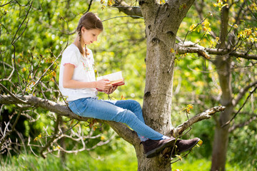 Young girl in white T-shirt sitting on the tree in the forest and reading interesting book. Great idea and activity to spend summer holidays in nature.