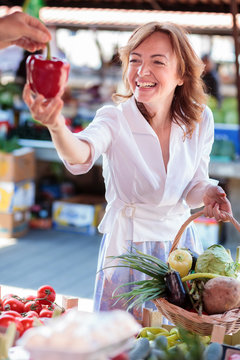 Happy mid adult Caucasian woman choosing and buying fresh organic vegetables in outdoor farmer's market, carrying shopping basket filled with vegetables
