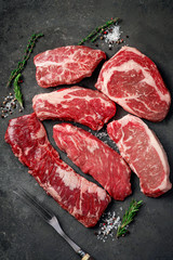 A set of different types of raw beef steaks: rib eye, Striploin, Denver, machete, top blade on a dark stone background.