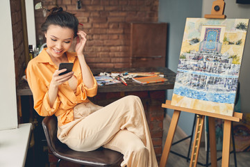 Cheerful female artist using cellphone in home art studio