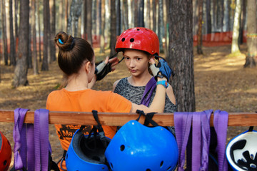 In a rope park, a beautiful young girl puts on equipment for traveling on a rope road, an instructor helps to put on a helmet, in a pine forest