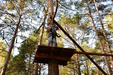 In a rope park, a beautiful young girl stands at high altitude, on a recreation area, against the backdrop of huge trees and a clear sky, in a pine forest