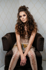 Portrait of young lady with dark long curly hair looking in camera. Girl sitting in a brown armchair.