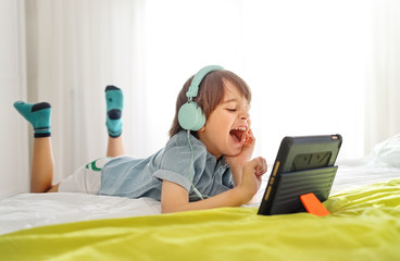 Little smiling boy siting on bed and playing on a digital tablet at home. Kid in his bedroom wearing headphones and using smart devices having fun online with his friend in a video call