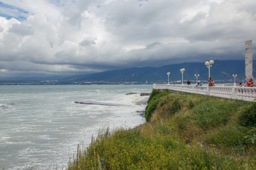 Storm in Gelendzhik Bay. Beach of Gelendzhik resort. Storm waves roll down a low stone pier located on the beach. People look at the waves from the embankment