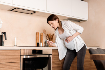 pregnant woman touching belly and having cramp while holding smartphone in kitchen