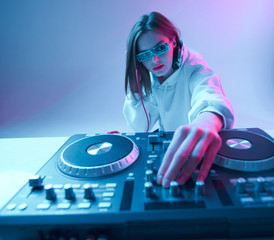 Cool young girl DJ mixes music on a mixing console and headphones, in stylish clothes, glasses on a...