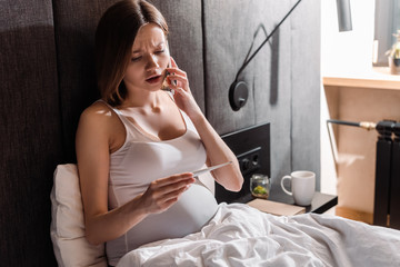 young pregnant woman talking on smartphone and looking at digital thermometer in bedroom
