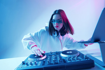 Cool young girl DJ mixes music on a mixing console and laptop, in stylish clothes, glasses on a...