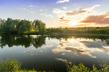 Fototapeta na wymiar Scenic view at beautiful spring sunset on a shiny lake with green branches, birch trees, bushes, grass, golden sun rays, calm water ,deep blue cloudy sky and forest on a background, spring landscape
