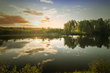 Fototapeta na wymiar Scenic view at beautiful spring sunset on a shiny lake with green branches, birch trees, bushes, grass, golden sun rays, calm water ,deep blue cloudy sky and forest on a background, spring landscape