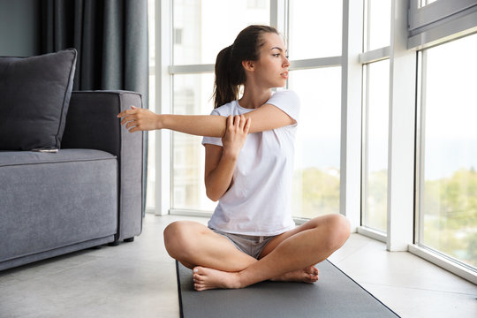 Image of focused young woman stretching her arm while sitting on yoga