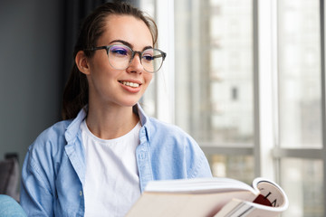 Image of cheerful woman reading book and smiling while sitting